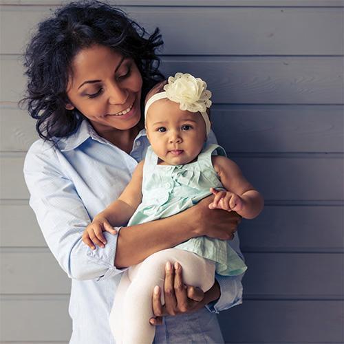 A woman casually dressed in a light blue button down shirt is looking down at the baby she's holding, the baby is wearing a light green dress and has on a headband with a flower