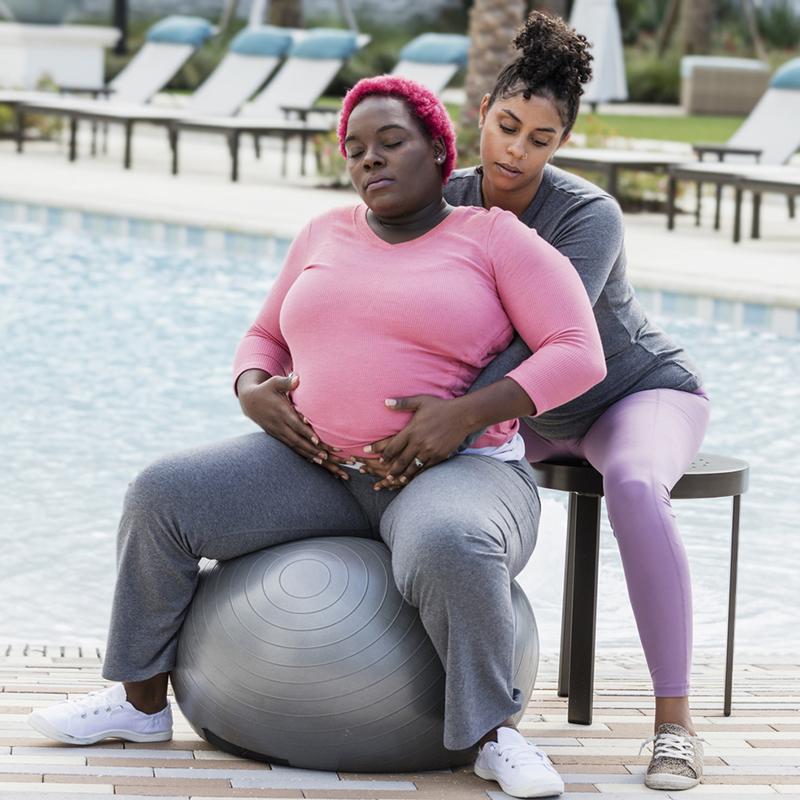 A pregnant Black woman with short, natural hair dyed fluorescent pink, wearing a pink long-sleeve workout top and gray yoga pants, is sitting on a gray yoga ball. A Black woman doula is sitting behind her and has her arms wrapped around the pregnant woman's stomach.