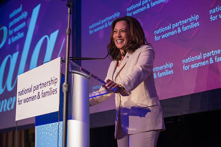 Kamala Harris stands on a stage at a podium with a National Partnership for Women and Families sign. The camera looks up at her as she smiles.