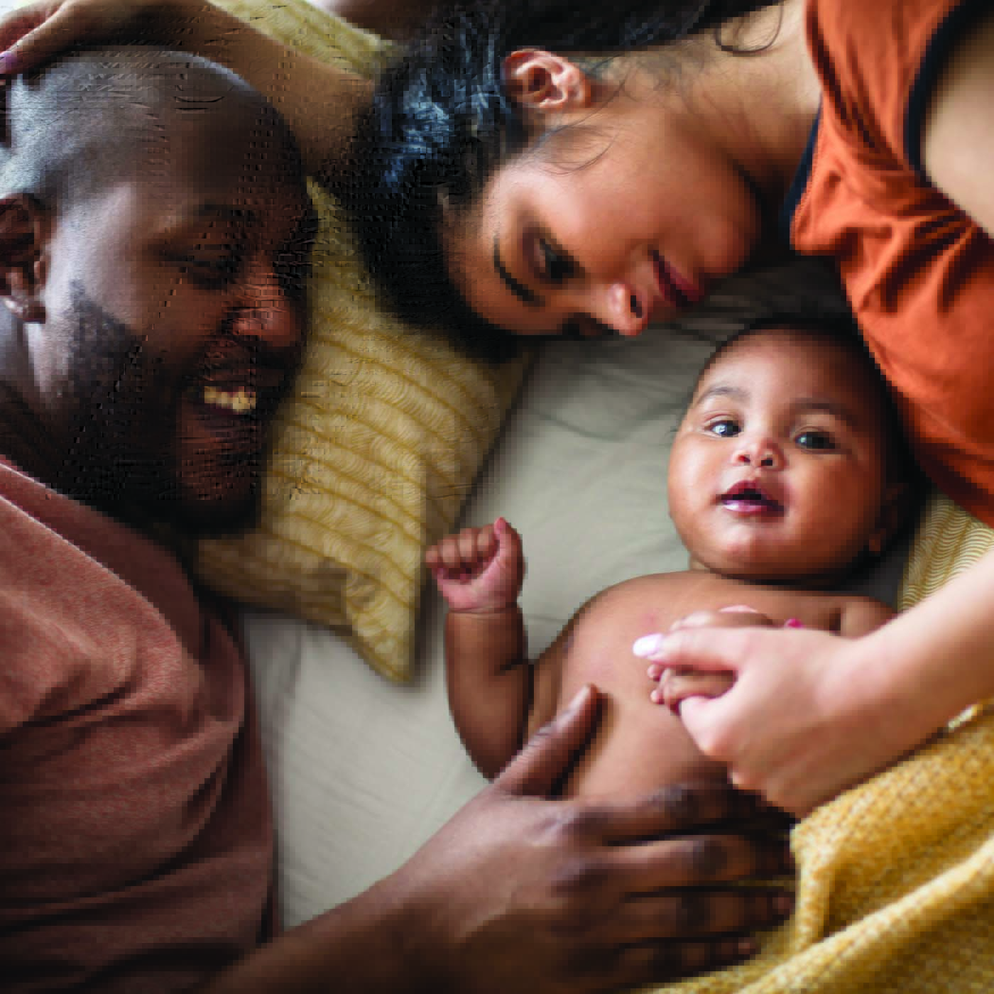 Parents who are lying down in bed, their heads on a pillow, are looking at and smiling at their infant child lying between them. The mother is holding the infant's hand and the father has his hand on the infant's belly.