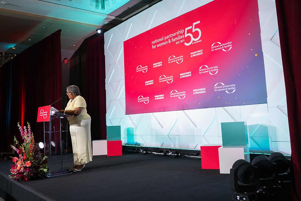 NPWF President Jocelyn Frye on stage behind at a podium with a large red step and repeat banner featuring the NPWF logo and theme Persistence & Progress at the 2026 Annual Event