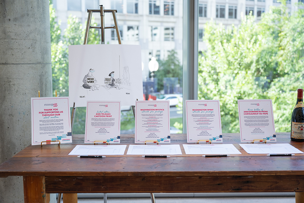 The 2026 Annual Event silent auction table with signs featuring items to bid on including an autographed cartoon print, sporting event tickets, and wine bottles