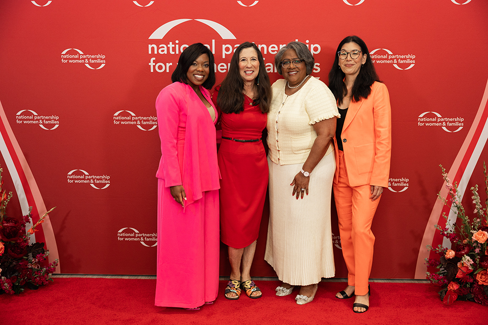 2026 Annual Event speakers from left to right: Del. Briana Sewell, Rep. Teresa Leger Fernández, Jocelyn Frye and Ai-jen Poo posing together on a red carpet in front of a red background with the National Partnership logo