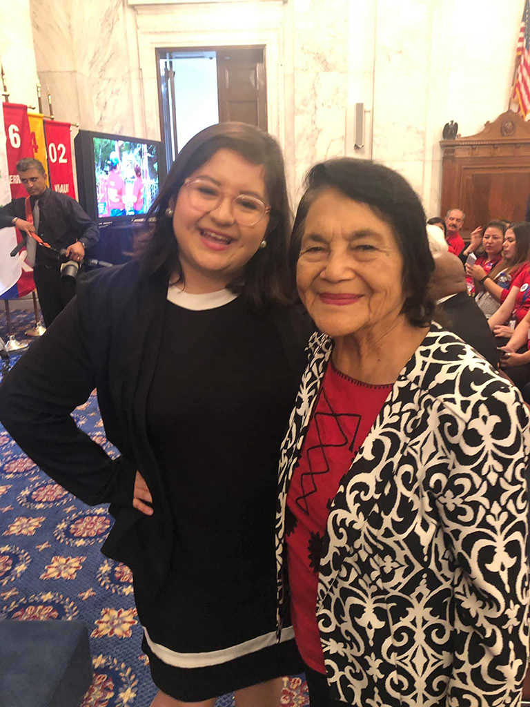 Belen Bonilla smiling and posing for a photo with Dolores Huerta who is also smiling, they are in the U.S. Senate's Kennedy Caucus Room and there is an American flag on display in the background