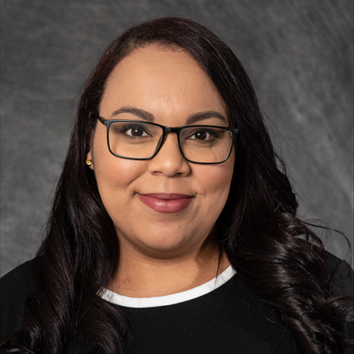 Portrait photo of Brittany Williams. She has long black hair that's slightly curled, is wearing black rimmed glasses, and a black dress.
