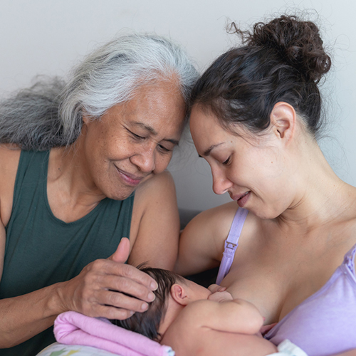 A happy young mother breastfeeds her infant daughter on a couch in a modern living room. Her mother is sitting next to her and lovingly caressing her granddaughter's head. The two adults' foreheads are leaned in and touching each other and both adults are looking lovingly at the infant.