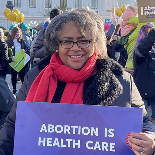 An image collage featuring protest signs in support of abortion rights and abortion access in front of the Supreme Court. The focal image is of Jocelyn Frye, National Partnership president, wearing a red scarf and a black coat, holding a sign that reads Abortion Is Health Care.