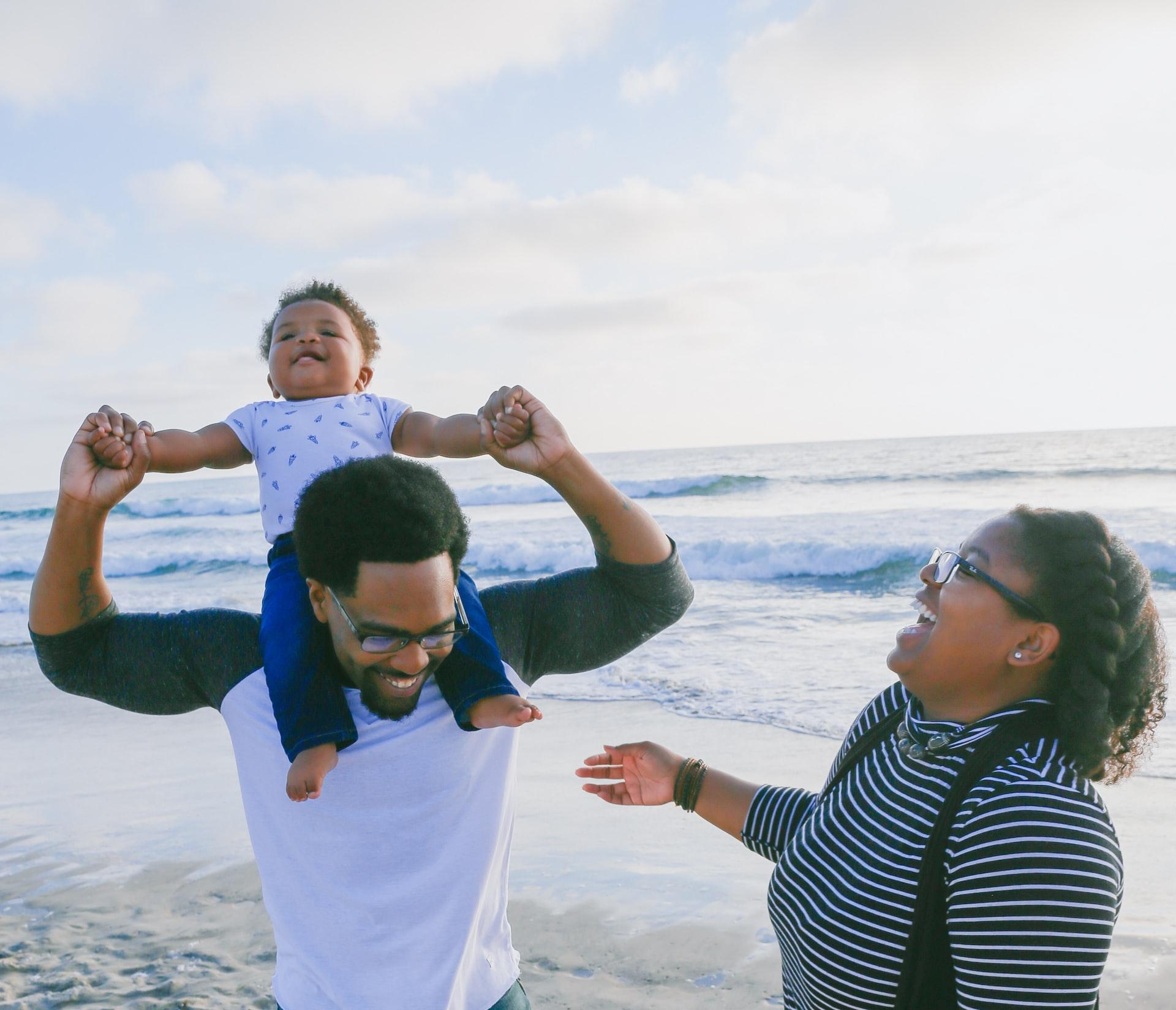 A Black man and his wife, a Black woman, are laughing as they're playing with their toddler who is sitting on the man's shoulders with the man holding the toddler's hands. They are at the beach, it's a sunny day with clouds in the sky and the shoreline is visible in the background.