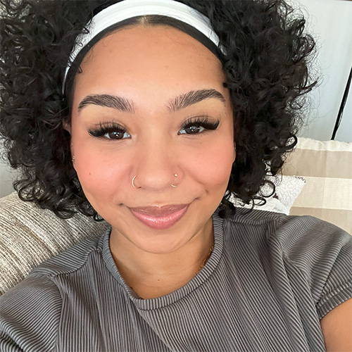 Portrait photo of Madison Alvarez. She is a young Afro-Latino woman with shoulder length black curly hair wearing a white headband and a gray stripped t-shirt.