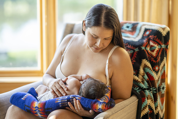 A beautiful photo of a young indigenous mother breastfeeding her newborn child. She is sitting on a comfortable chair in a living room at home next to big bright windows. She is lovingly looking at her baby daughter.