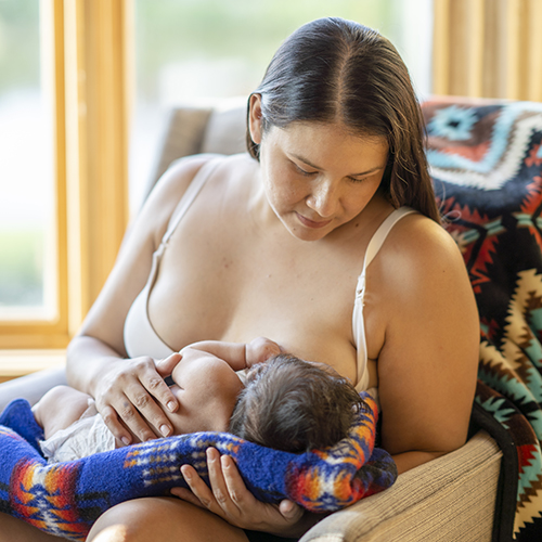 A beautiful photo of a young indigenous mother breastfeeding her newborn child. She is sitting on a comfortable chair in a living room at home next to big bright windows. She is lovingly looking at her baby daughter.