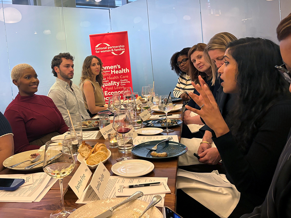 8 participants are sitting around a long wood dining table, all are listening intently to one woman who's speaking and gesturing with her hands.