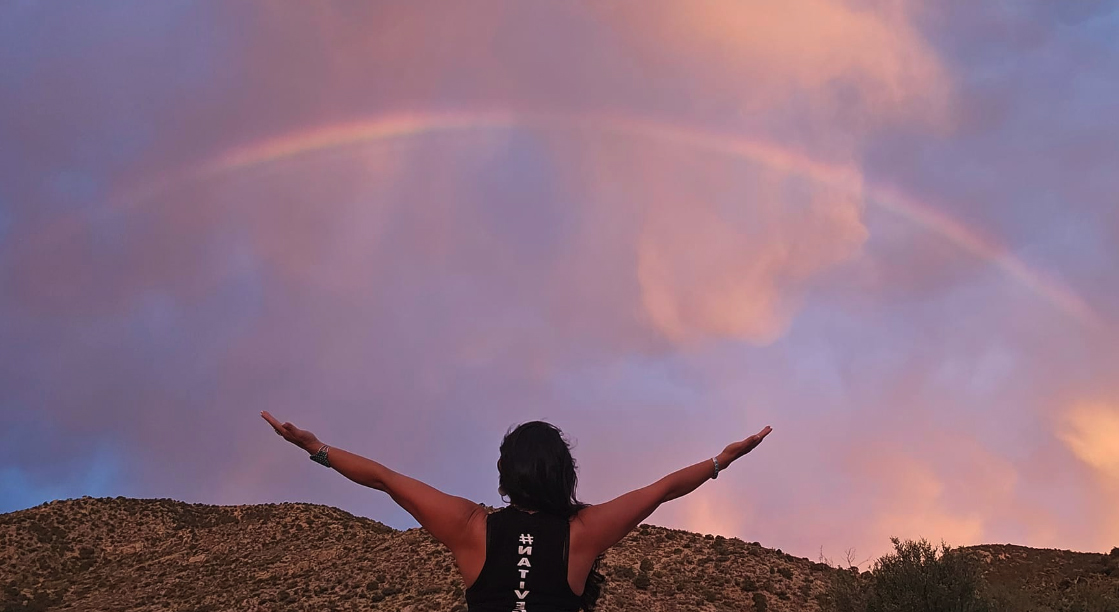 A Native woman in a desert, she is facing away from the camera toward the pink-blue sky with her arms outstretched