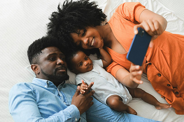 A Black family -- a Black man and Black woman -- taking a selfie photo with their infant cuddled in between them. The woman is holding up a smartphone and using the screen to compose the photo. She's wearing an orange dress. The dad is holding his baby's right hand and he's wearing a light blue denim button-up.