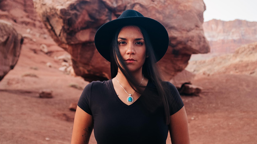 A Native woman standing in a dessert area with natural rock formations. She has long straight dark hair, and is wearing a black v-neck t-shirt, a silver necklace with a turquoise pendant, and a black rimmed hat.