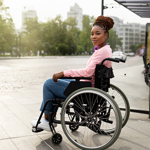 A Black woman in a wheelchair with braids in a bun waits for a bus by a bus shelter. Her head is turned to the left, she's smiling slightly and looking directly into the camera.