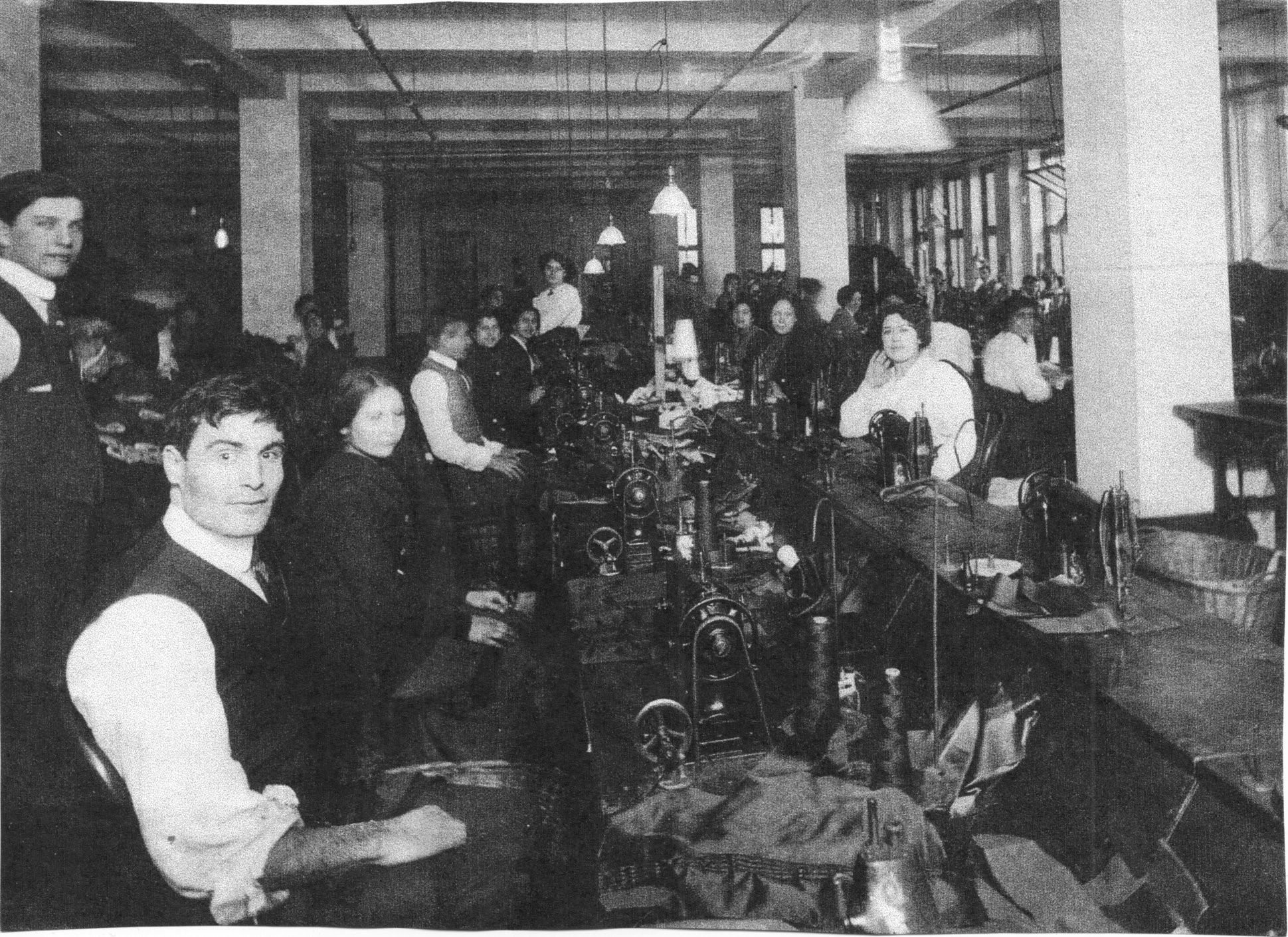 Photo description: Men and women working at the Triangle Shirtwaist Factory, some are standing and some are seated. Most are looking toward the camera. There are eight people seated at either side of a long table that's set up for tactile factory work – in front of each seated person is a sewing machine. In the foreground on the left is a seated man with short dark hair, he's wearing a button-up vest with a white button down shirt with the sleeves rolled up. To his left is a young woman, also seated, in a dark coat and long skirt, her hair is tied back in a low bun. In the midground, on the right side of the photo, is a young woman in a white shirt. She is seated and facing the camera, her right arm is propped up on the table and her right hand is touching her face. She has short, curly hair. This is the author, Marissa Ditowsky's, great grandmother.